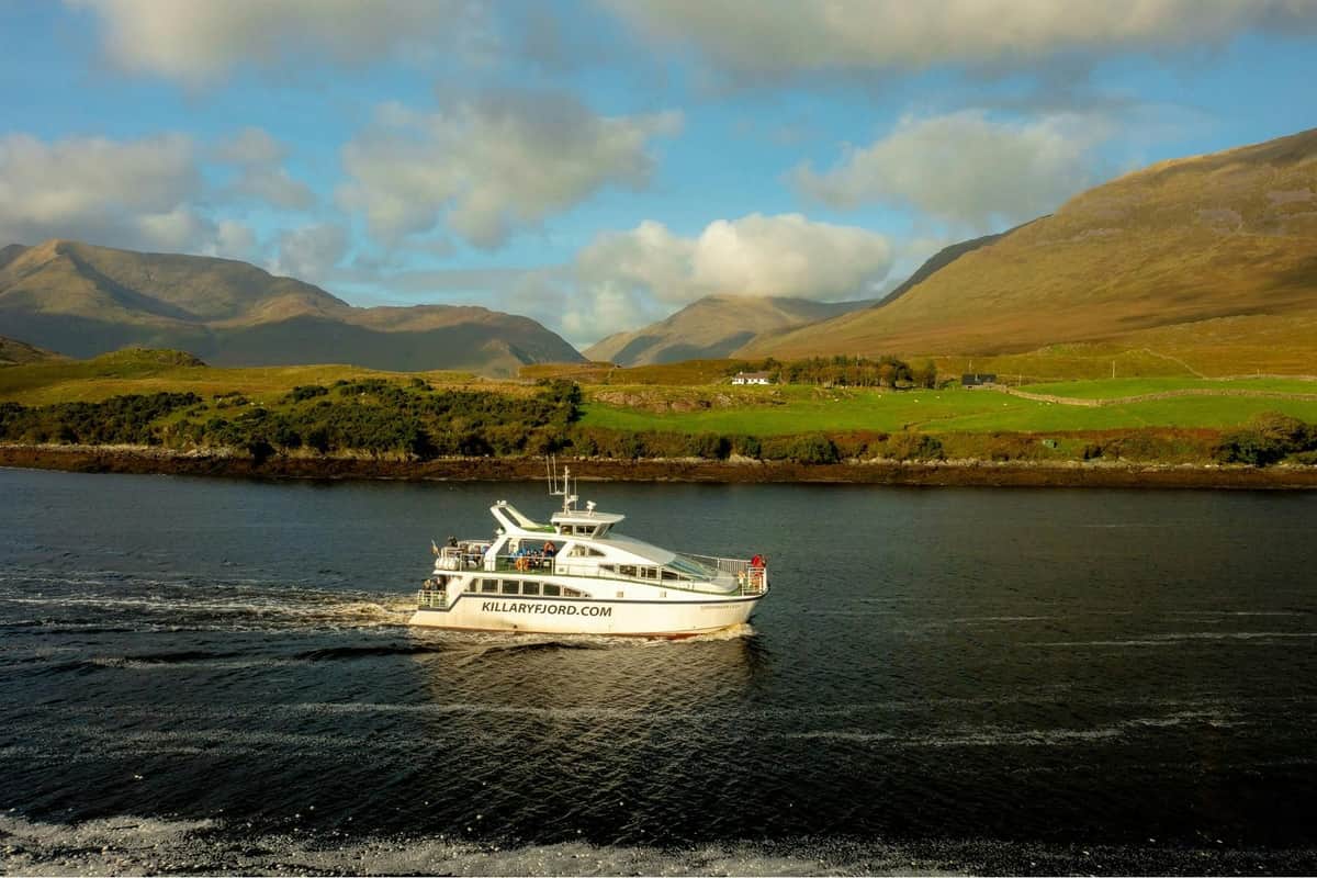 Un barco con la etiqueta "killaryfjord.com" navega por una estrecha masa de agua, con verdes colinas y montañas al fondo.