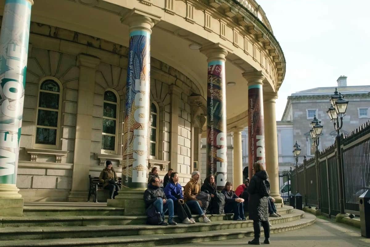 Dublin: Guided Dark Walking Tour People sitting and talking on the steps of a building with large columns featuring the words "EXPLORE," "READ," and "LEARN."