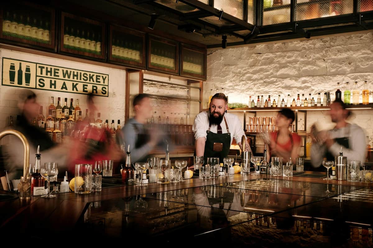 A bartender with a beard stands behind a bar, surrounded by four blurred people amidst a variety of glasses and bottles.