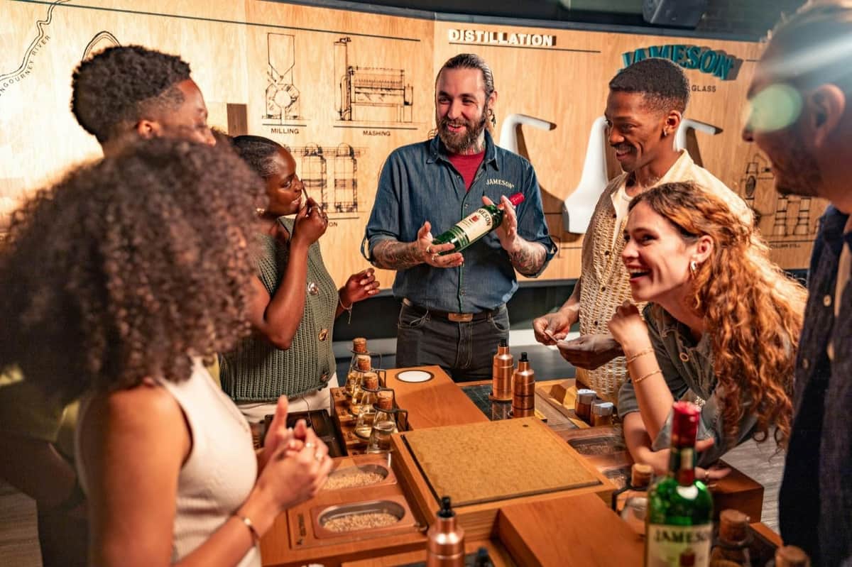 A group of people enjoying a whiskey-tasting session at a distillery, with a presenter holding a bottle of Jameson.