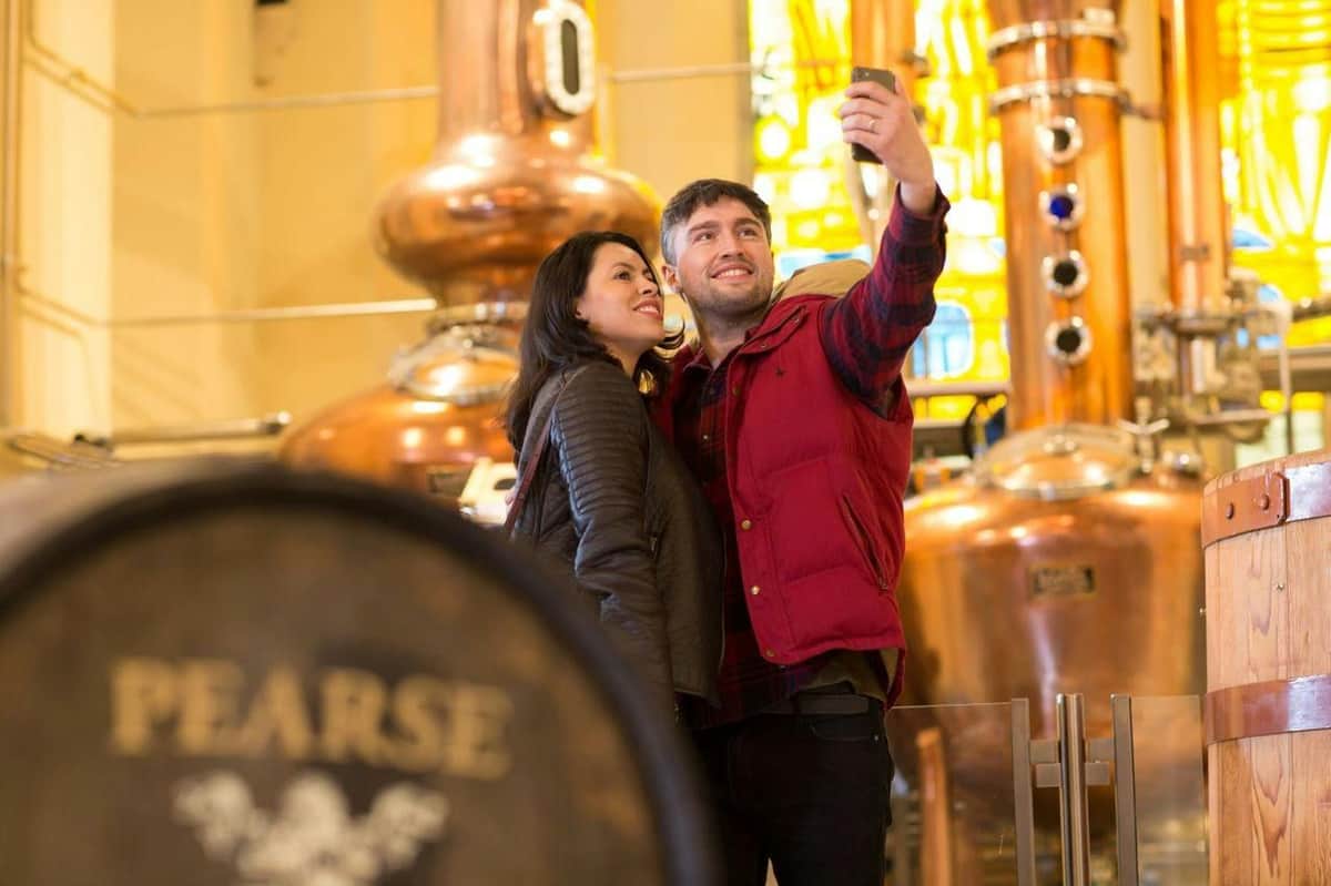 A smiling couple takes a selfie in front of large copper distillery tanks with stained glass in the background.
