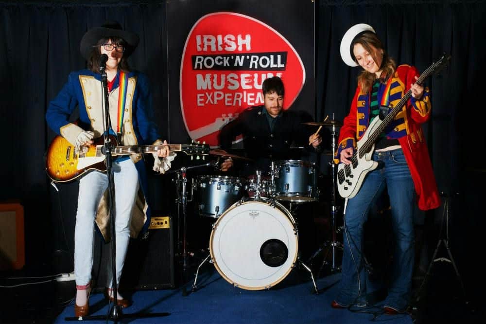 Une personne vêtue d'une tenue colorée, semblable à celle d'un groupe, joue d'une guitare basse blanche à côté d'un batteur, avec une bannière "Irish Rock 'N' Roll Museum Experience" derrière elle.