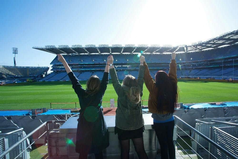 Three individuals with raised arms stand facing a large, empty stadium under bright sunlight.