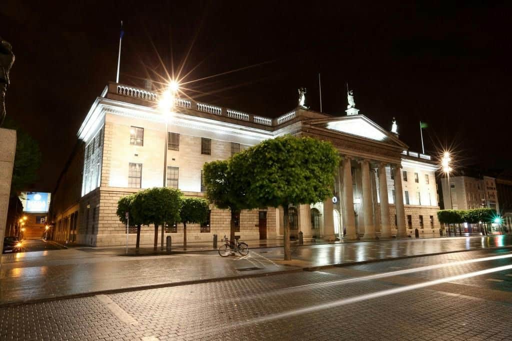 General Post Office Dublin Illuminated neoclassical building with columns and statues at night, surrounded by trees and a wet pavement reflecting lights.
