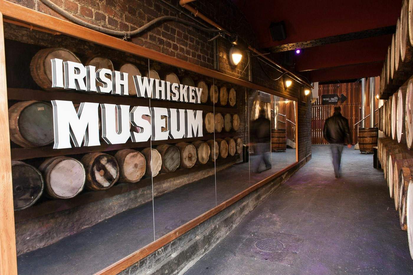 Irish Whiskey Museum Dimly-lit hallway with whiskey barrels on both sides and a sign that says "Irish Whiskey Museum." A person walks into the distance.