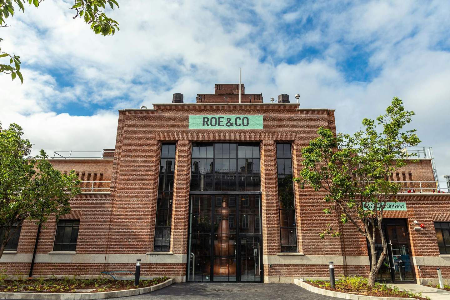 Brick building with large windows, “ROE & CO” sign, trees in the foreground, and a clear blue sky with some clouds.