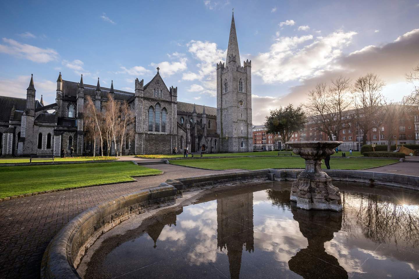 Saint Patrick's Cathedral A historic stone cathedral with a tall spire beside a reflective fountain, surrounded by green lawns and a few people under a blue sky.