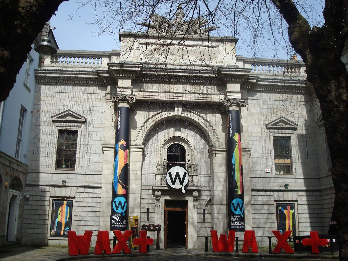 Façade du bâtiment du musée de la cire avec un extérieur en pierre, de grands piliers, des bannières et un panneau "WAX+".