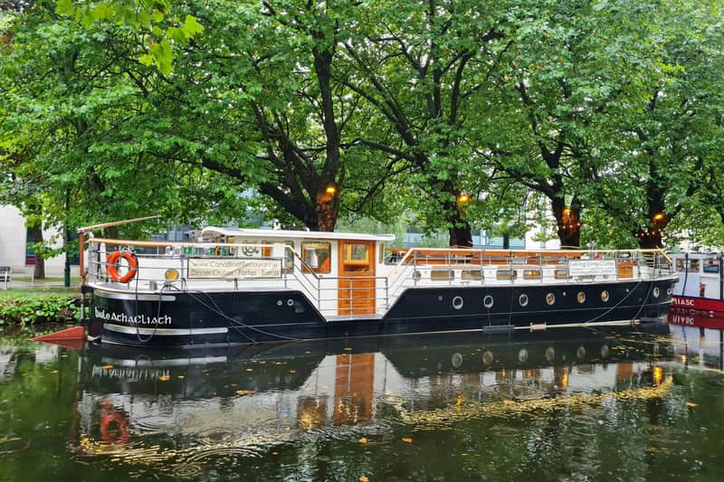 A moored canal boat with black and white exterior and wooden details, set against lush green trees and calm water.
