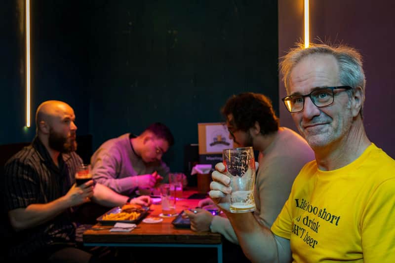 Dublin: Small Group Guided Craft Beer Walking Tour + Lunch A man in a yellow shirt holding a glass in a dimly lit room with people sitting at a table in the background.