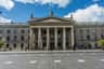 Historic building with large columns and statues on the roof, an Irish flag, and people walking in front on a stone plaza.